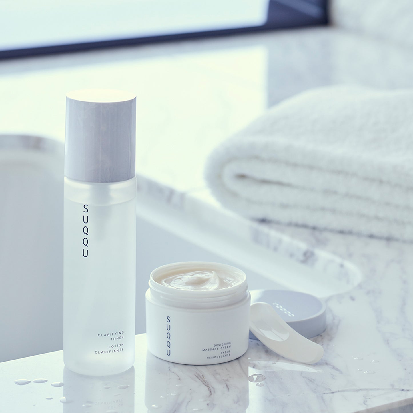 SUQQU Designing Massage Cream tub with lid open showing cream on bathroom table with other skincare products.