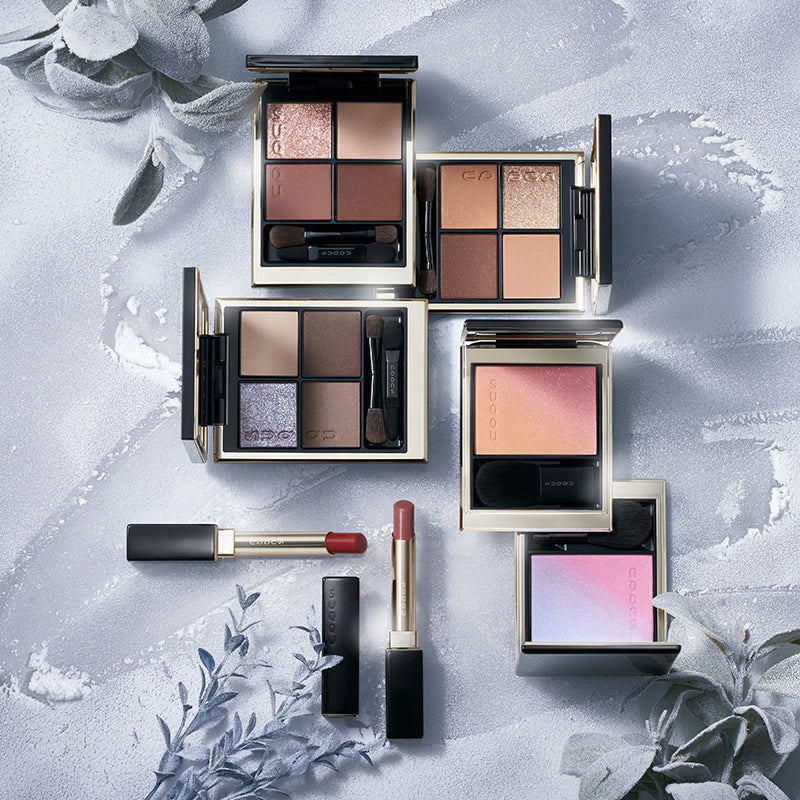 SUQQU Winter Colour Collection 2025 product group on frosted plant background. Multiple makeup palettes and lipsticks laid out together.