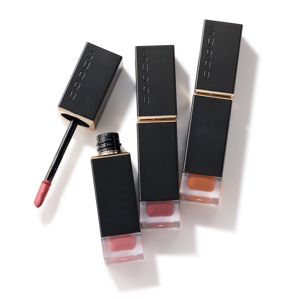 Three SUQQU Comfort Lip Fluid Fog make up products on plain background with one case open showing liquid lip colour wand.