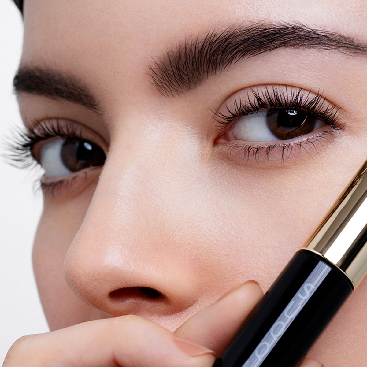 Close-up of a woman's face holding a black and gold SUQQU 3D Mascara near her eye, showing detailed lengthened, thickened and airy fanned out eyelashes and smooth skin against a light background.