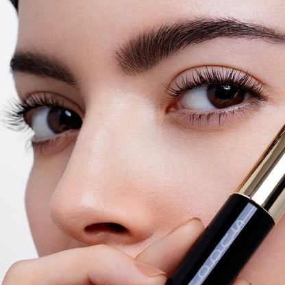 Close-up of a woman's face holding a black and gold SUQQU 3D Mascara near her eye, showing detailed lengthened, thickened and airy fanned out eyelashes and smooth skin against a light background.