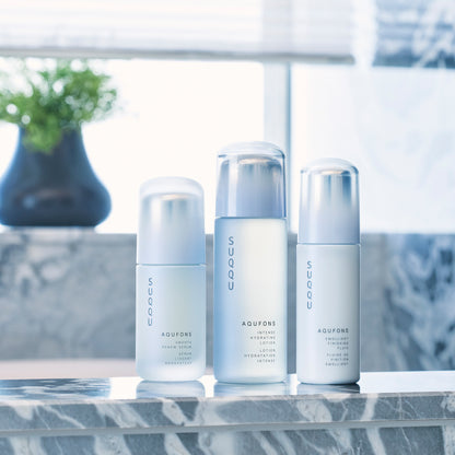 Three skincare products from SUQQU's 'Aqufons' range standing in a line on a bathroom table. Serum, lotion and fluid face moisturisers in pump bottles.