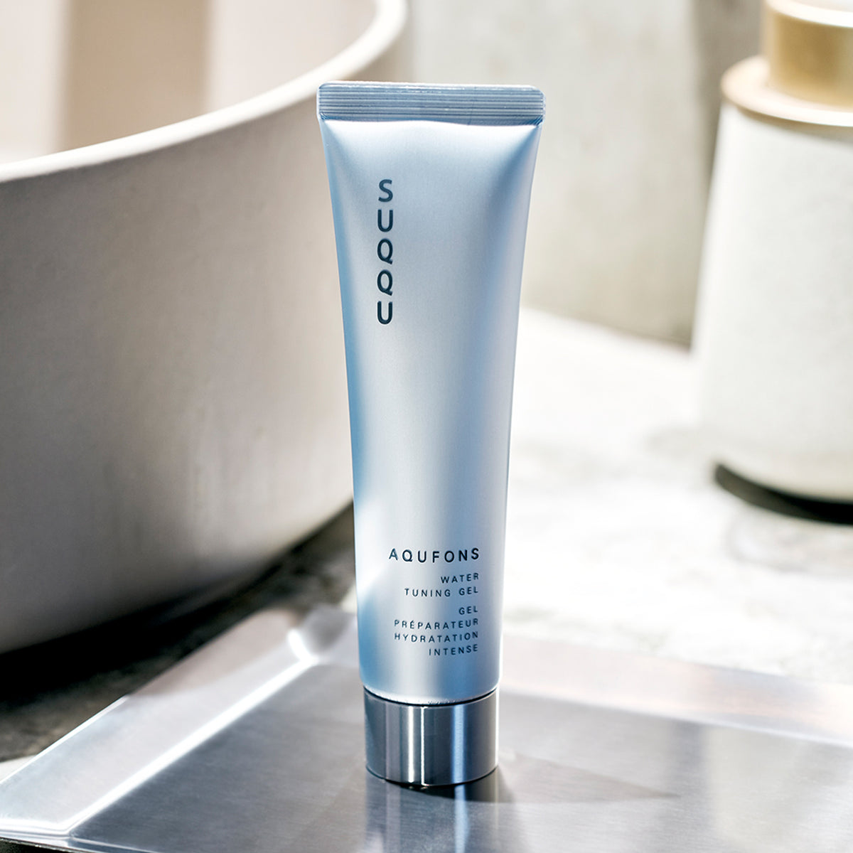 A tube of SUQQU Aquafons Water Tuning Gel standing on a metallic tray in a bathroom setting with a bathtub and toiletry container blurred in the background.