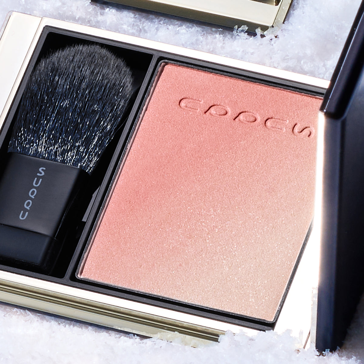SUQQU Blurring Color Blush 119 open black and gold luxury blusher compact on snowy background. Coral pink and pale beige pink highlighter shades.