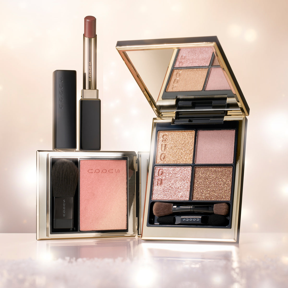 SUQQU Winter Color Collection 2026 cosmetics makeup set featuring a dark reddish pink lipstick, a square compact with a pink blush and brush, and an open square four-pan eyeshadow palette with gold and pink shades.