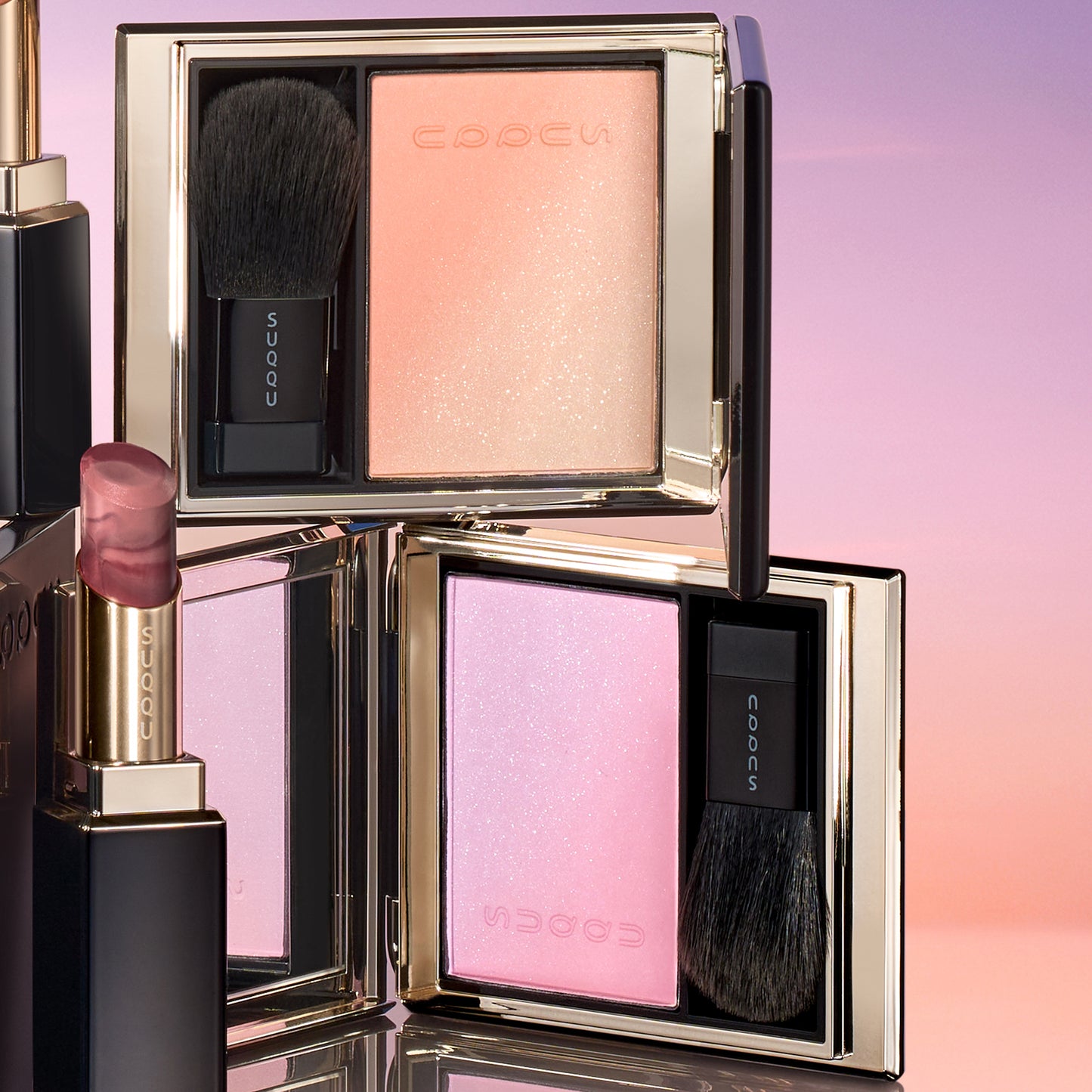 SUQQU 2026 Summer Collection, multiple beautiful colourful luxury makeup products on a gradient sky background. Two blushers with orange gold and lilac pink and two lipsticks in orange red and grape red.