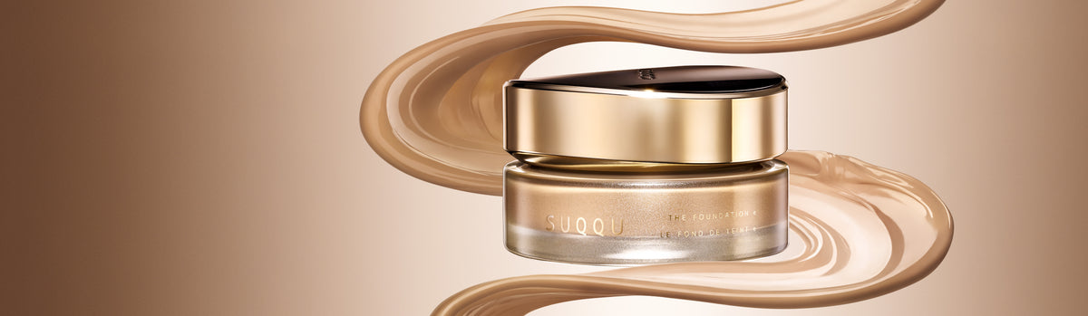 SUQQU 'The Foundation e' jar with a black lid and metallic rose-gold base, surrounded by flowing beige foundation swirled with a cream background.