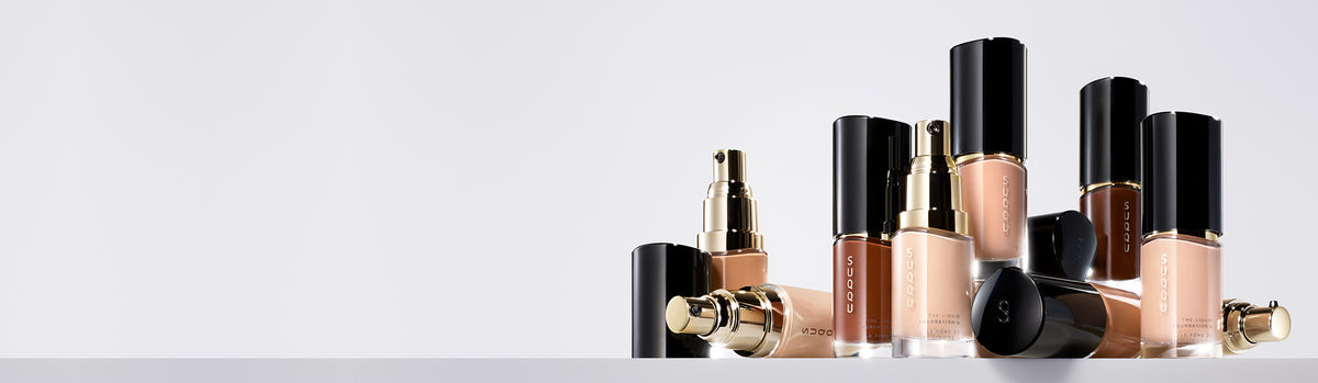 Multiple luxurious SUQQU 'The Liquid Foundation e' bottles standing on a table in various shades.