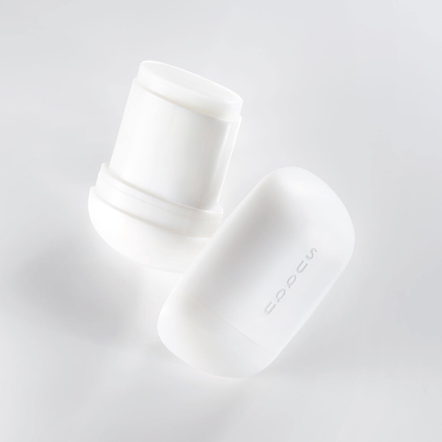 Two SUQQU Glowing Balm products floating on a soft grey background, one is open showing balm stick, the other is closed.