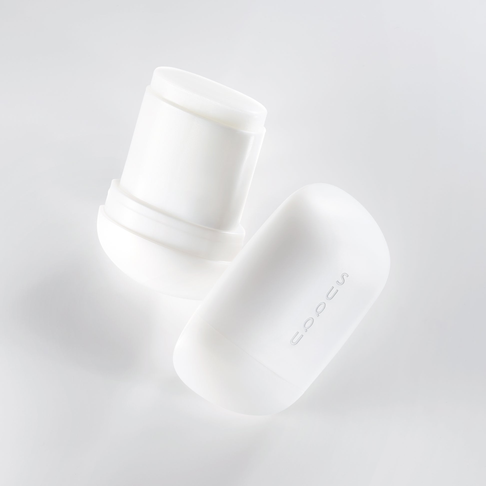 Two SUQQU Glowing Balm products floating on a soft grey background, one is open showing balm stick, the other is closed.