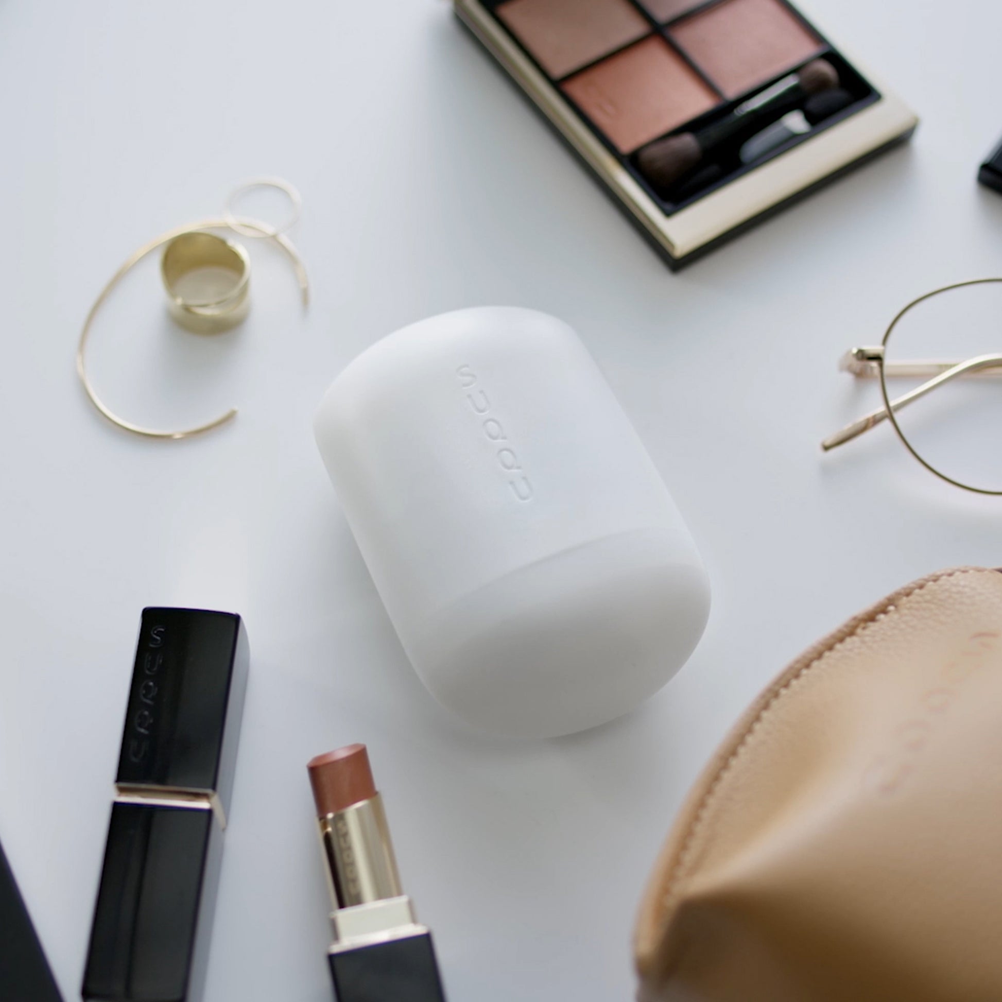 SUQQU Glowing Balm product on a table surrounded by other personal effects such as a bag, earrings, lipstick and glasses.