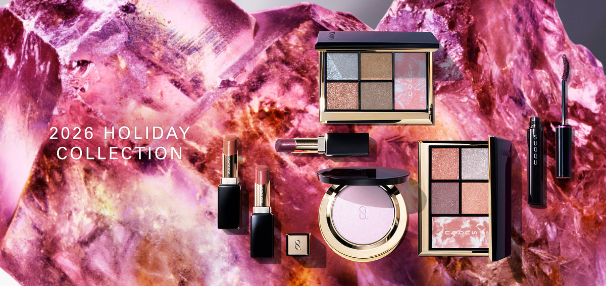 A flat-lay collection of SUQQU luxury Christmas Gift makeup products including eyeshadow and blusher combination palettes, lipsticks and mascara, arranged on a sparkling pink crystal background with the text '2026 HOLIDAY COLLECTION'.