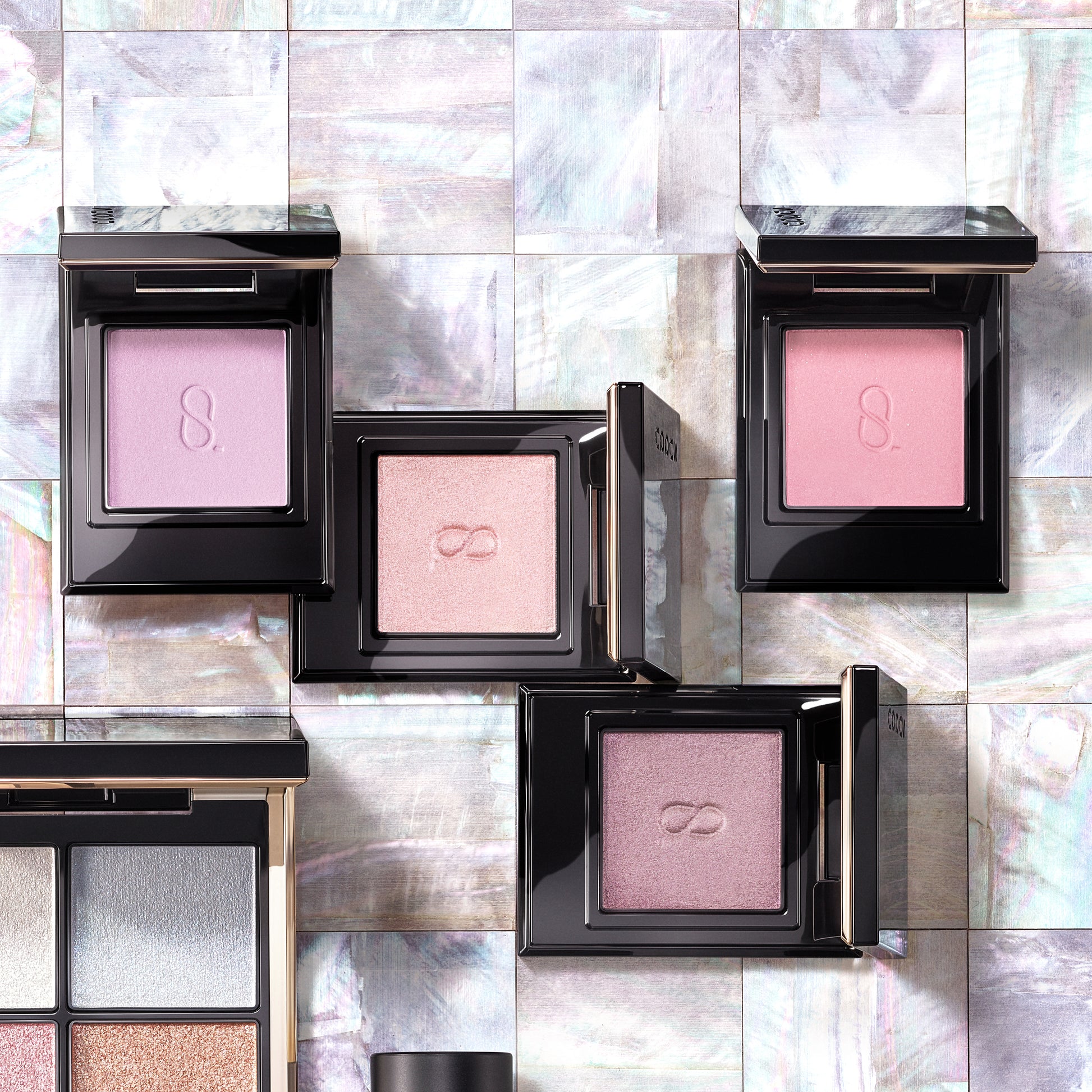 Four beautiful SUQQU Mono Look Eyes single luxury eyeshadow palettes open on a pearl tile table. In pale soft pink and purple, pearlescent glitter and matte finishes.