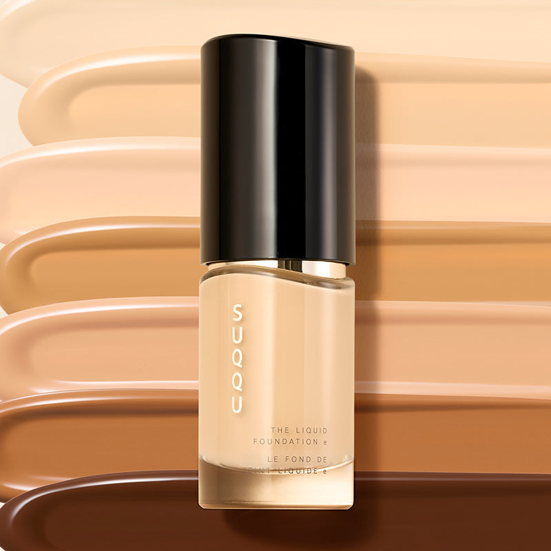 A pump bottle of SUQQU The Liquid Foundation e, featuring a black cap and clear bottle showing the beige foundation inside, set against a background of horizontal liquid foundation swatches in various shades.