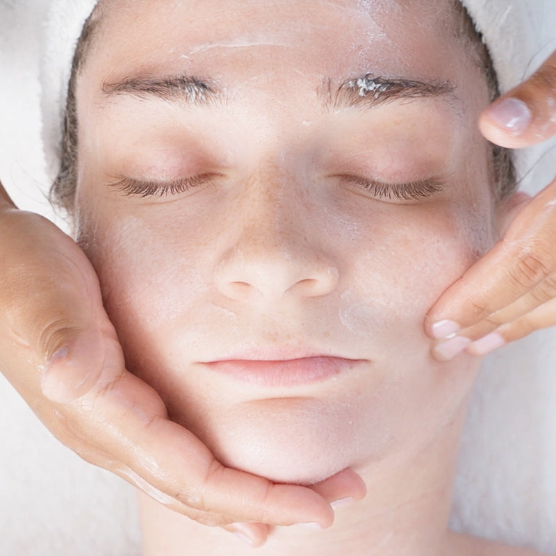 Close-up of a person receiving a GANKIN facial treatment, with hands gently holding the cheeks and forehead while a white head wrap is visible.