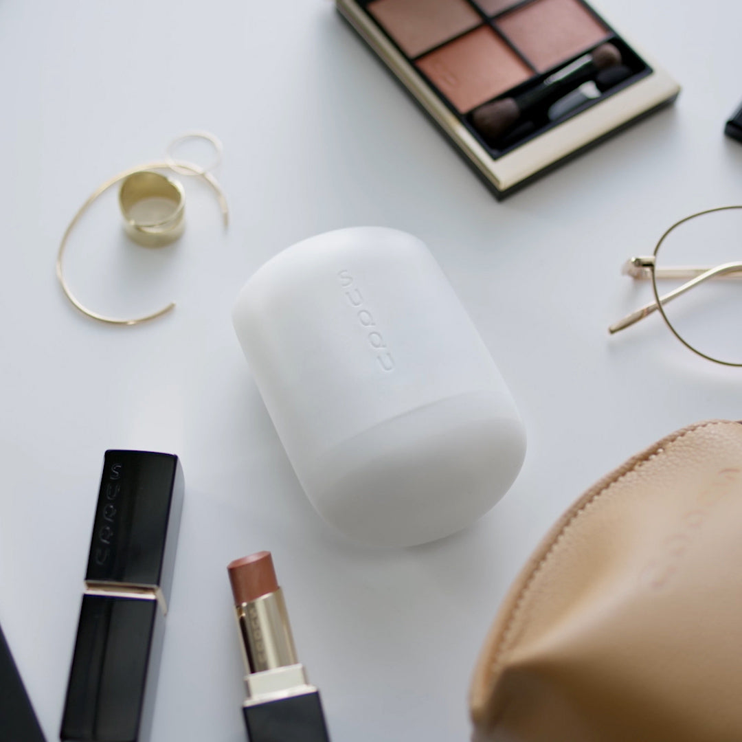 SUQQU Glowing Balm product on a table surrounded by other personal effects such as a bag, earrings, lipstick and glasses.