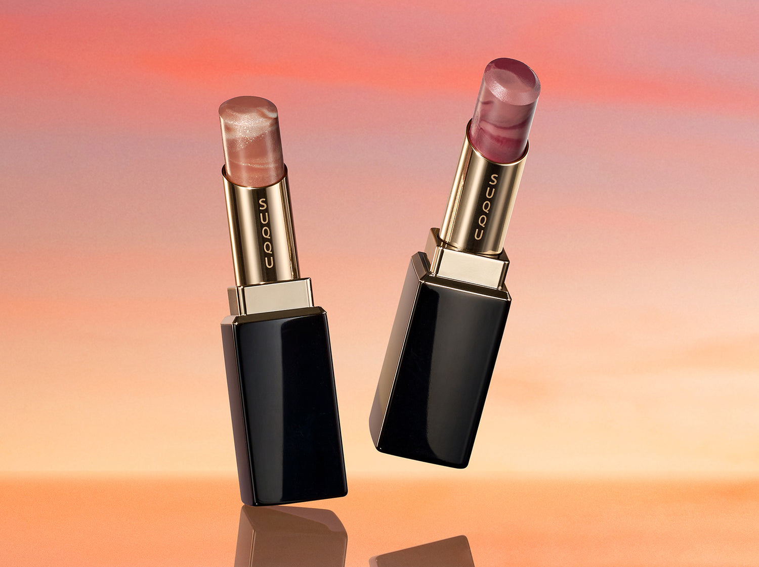 SUQQU 2026 Summer Collection Moisture Glaze Lipstick 112 and 113 open on a sunset sky background. One lipstick in a golden brown and one in a deep berry red. Discover some of the very the best lipsticks from Japan at SUQQU.