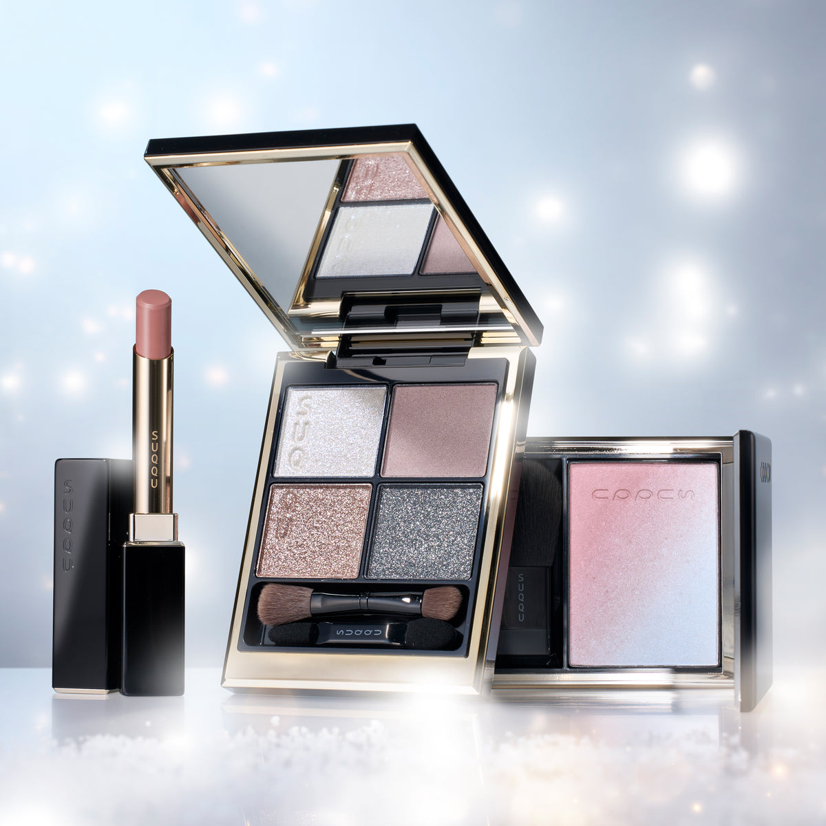 A SUQQU Winter Color Collection 2026  cosmetics set featuring an open four-shade eyeshadow palette with shimmering neutral and metallic shades, a lipstick in a black tube with a pink bullet, and a square highlighter or blush compact with a pink-to-blue gradient, all arranged on a reflective white surface against a soft light blue sparkling background.