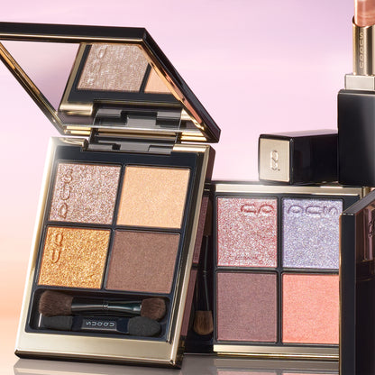 SUQQU 2026 Summer Collection, multiple beautiful colourful luxury makeup products on a gradient sky background. Two eyeshadows with gold and pink coral tones, with lipstick in orange red.
