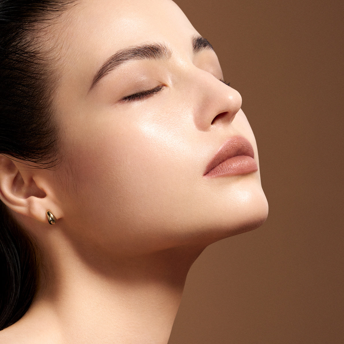 Close-up of a woman with flawless skin showcasing SUQQU 'The Foundation e' cream foundation base make up. With her eyes closed wearing a small gold hoop earring, set against a warm brown background.