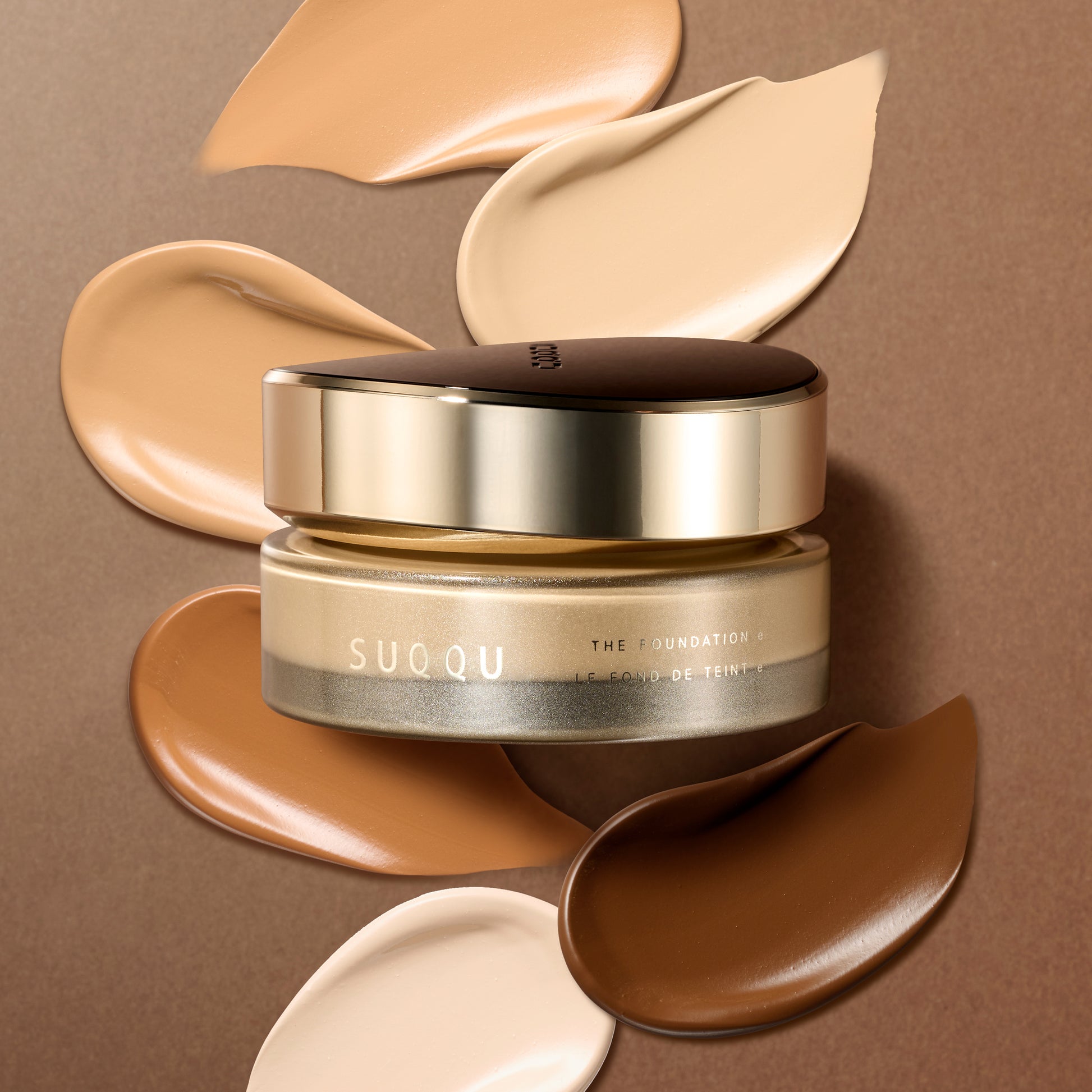 A jar of SUOQU The Foundation e on a brown background, surrounded by smooth and super creamy, swirled makeup swatches in various skin tones.