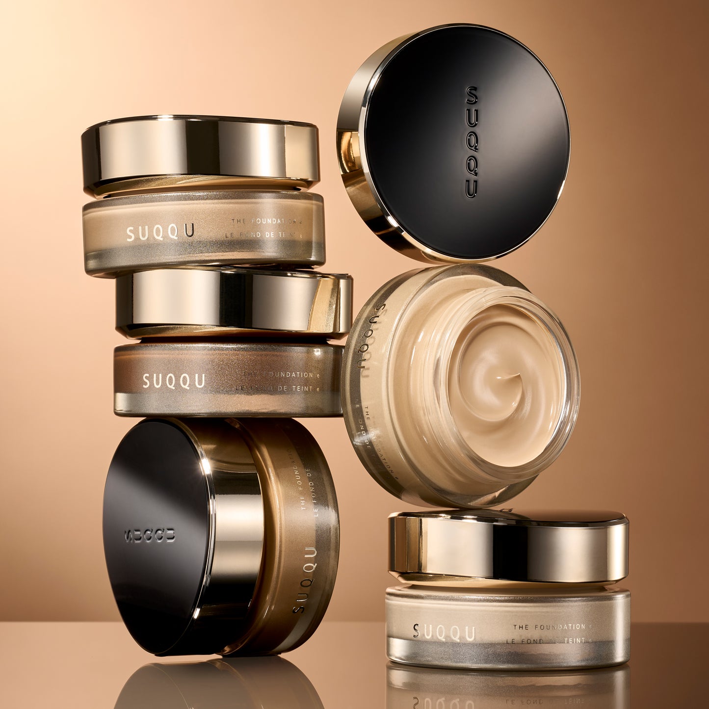 A group of stacked SUQQU 'The Foundation e' luxury base makeup jars  against a warm beige background. The products feature metallic gold and black lids with frosted glass containers showing beige and brown toned cream foundation inside.