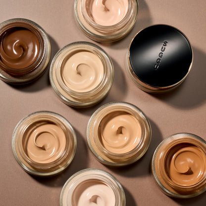 An overhead flat-lay of open cosmetic jars showing cream foundation makeup in multiple skin-tone shades, with one closed jar featuring a black lid printed with the brand name SUQQU.