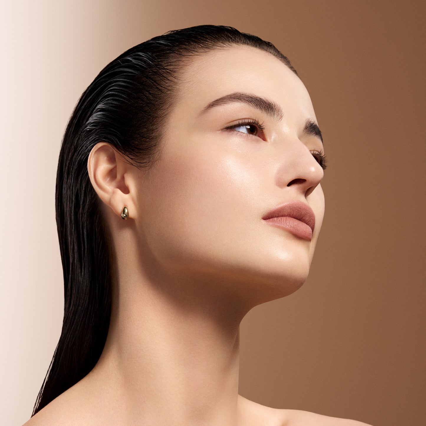 A close-up portrait of a woman with wet, slicked-back black hair wearing a small gold hoop earring, set against a warm beige and brown gradient background, showing skin with the flawless, smooth finish and clear radiant glow of SUQQU The Foundation e.