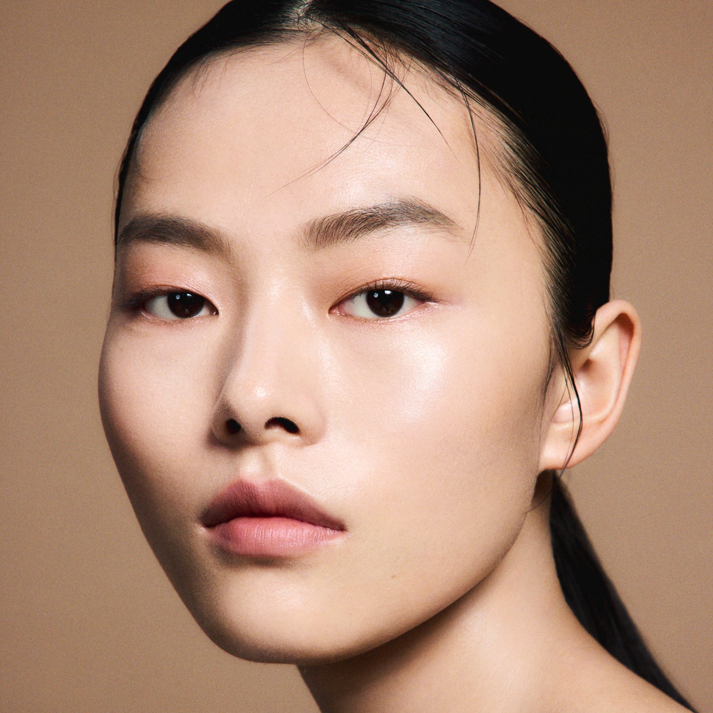 A close-up portrait of a woman with dark hair pulled back and flawlessly smooth and radiant skin, wearing SUQQU 'The Foundation e'  and a neutral expression against a warm beige background.