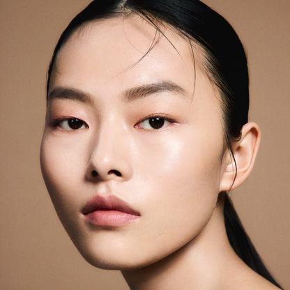 A close-up portrait of a woman with dark hair pulled back and flawlessly smooth and radiant skin, wearing SUQQU 'The Foundation e'  and a neutral expression against a warm beige background.