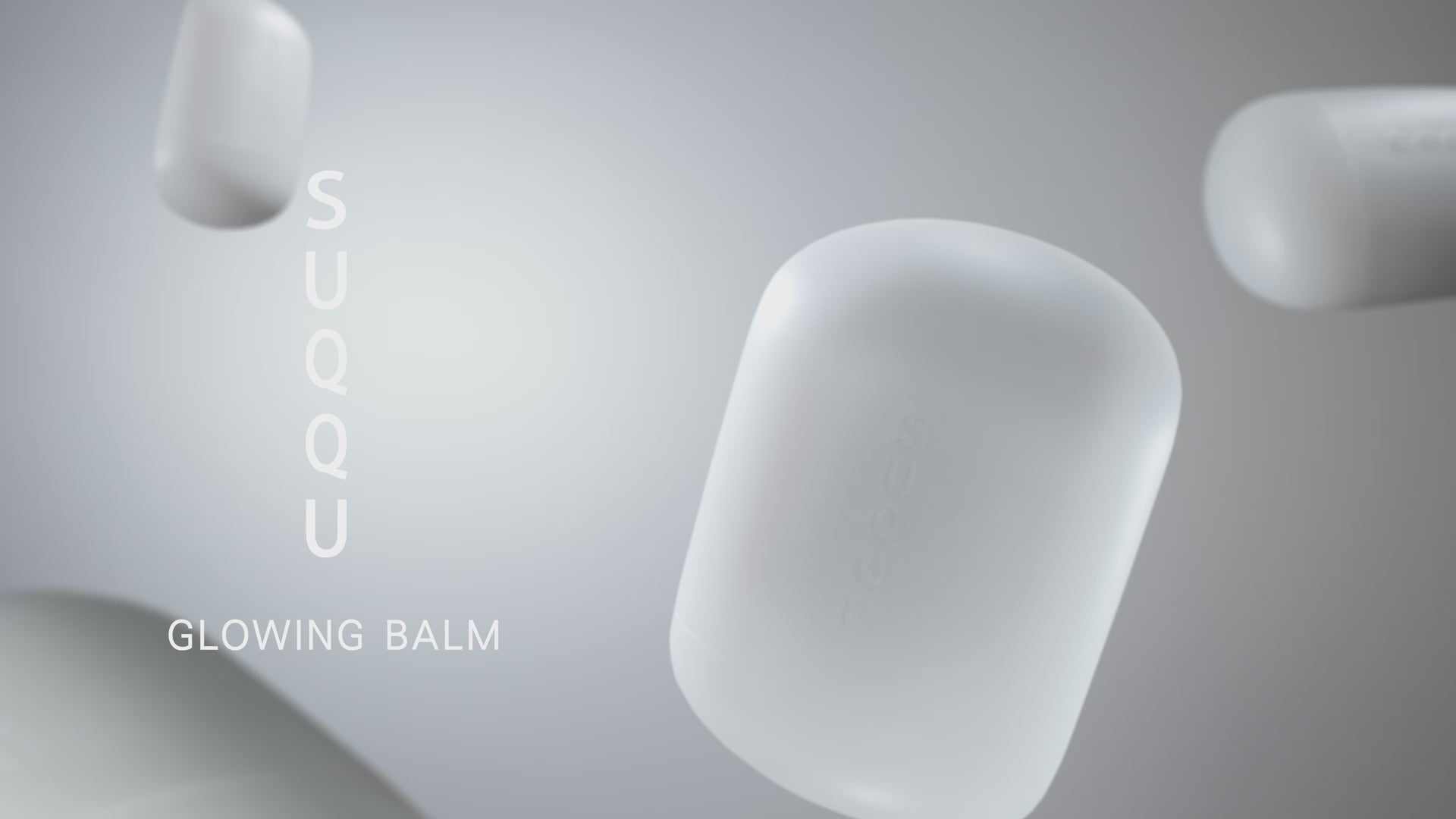 Load video: Multiple SUQQU Glowing Balm products floating and rotating. Product is shown on a table with other personal effects, then being open and applied to the back of the hand and face with a close up a model.
