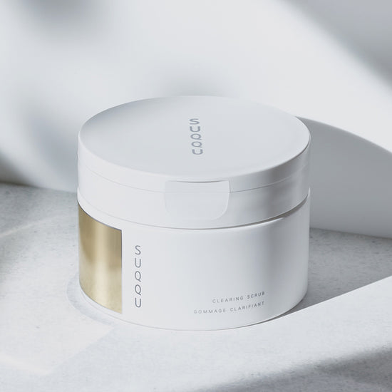 SUQQU Clearing Scrub tub shown as it is opened and the creamy texture is taken with a spoon, then applied to the back of a hand and massaged.