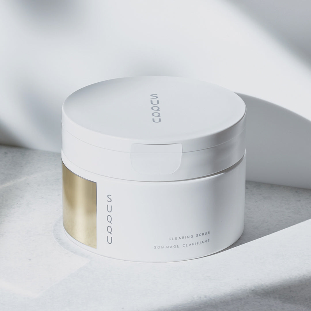 SUQQU Clearing Scrub tub shown as it is opened and the creamy texture is taken with a spoon, then applied to the back of a hand and massaged.