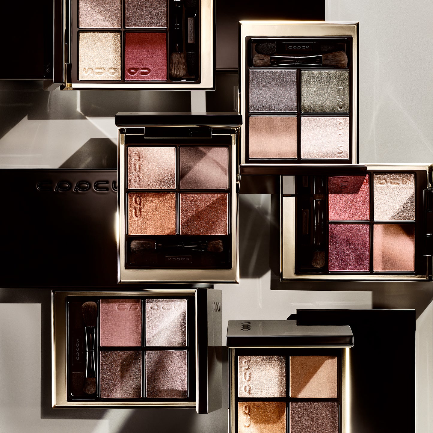 Group of stacked SUQQU Signature Color Eyes eyeshadow quad colour make up palettes open on plain background.
