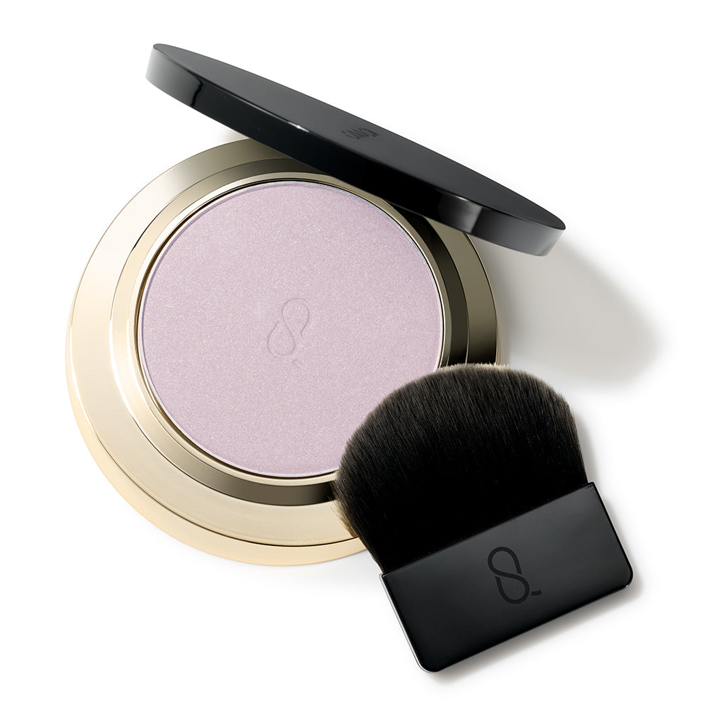 An open round SUQQU Glow Setting Powder e 101 face powder makeup compact with a black lid resting above the compact, showing a shimmering light lavender-pink pressed powder in a metallic gold case on a white background, shown with brush.